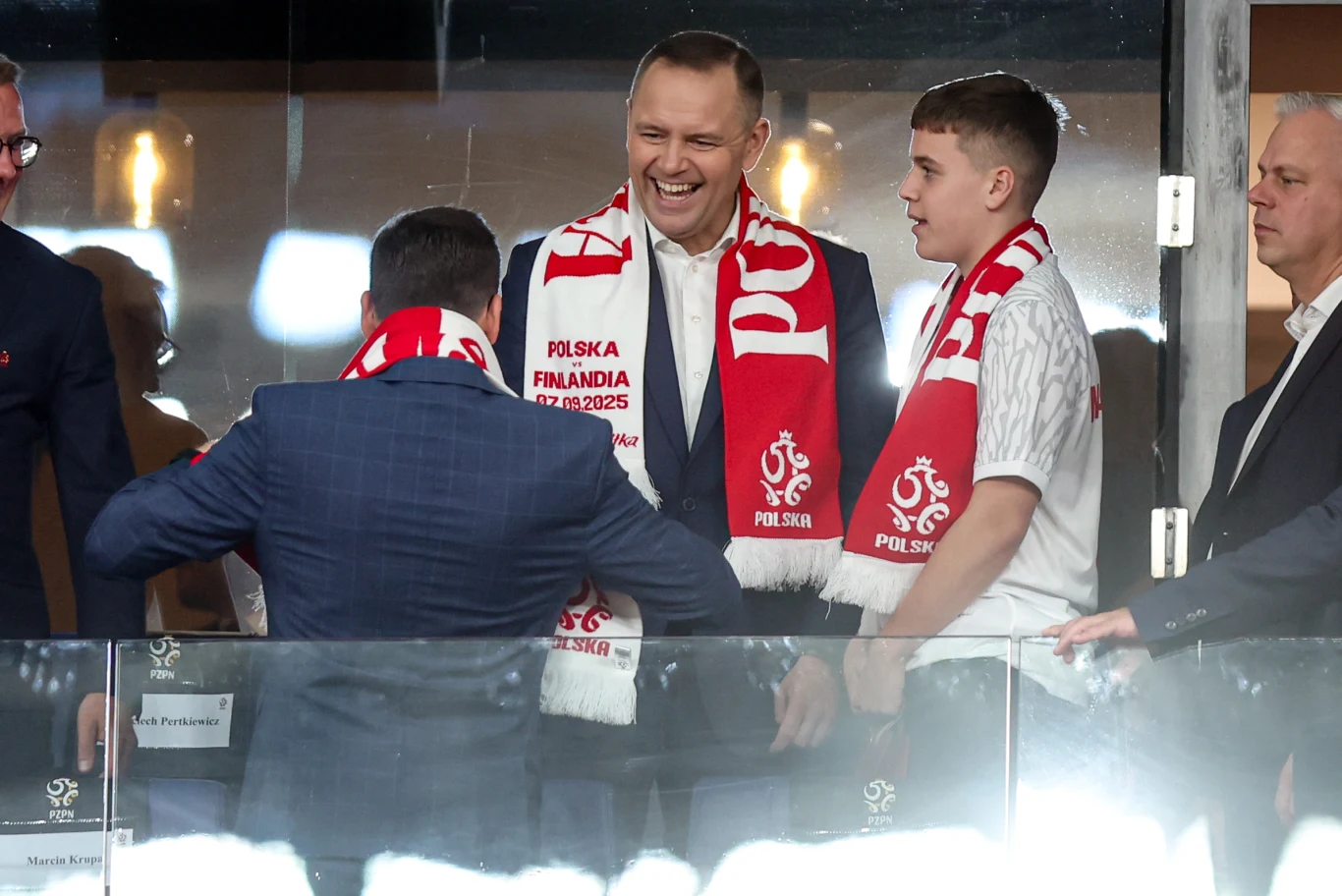 Czterech mężczyzn w otoczeniu przeszklonym, dwaj z nich uśmiechnięci z szalikami w barwach Polski z napisem 'POLSKA' i datą meczu Polska-Finlandia, stoją blisko siebie, prowadząc ożywioną rozmowę.