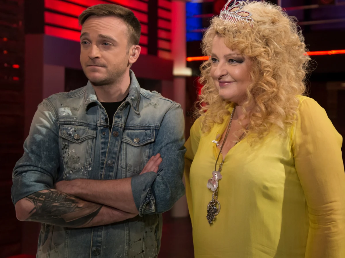 Mężczyzna w dżinsowej kurtce z tatuażem na przedramieniu stoi obok kobiety w żółtej bluzce z blond, kręconymi włosami i koroną na głowie, oboje patrzą w jednym kierunku w kolorowym, telewizyjnym studio.
