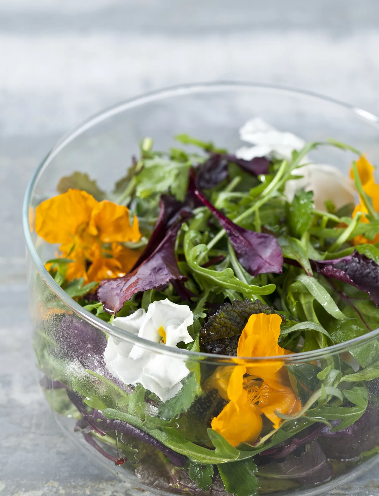 Miska pełna świeżych liści sałaty, mieszanki ziół i kolorowych, jadalnych kwiatów o intensywnie pomarańczowych i białych płatkach.