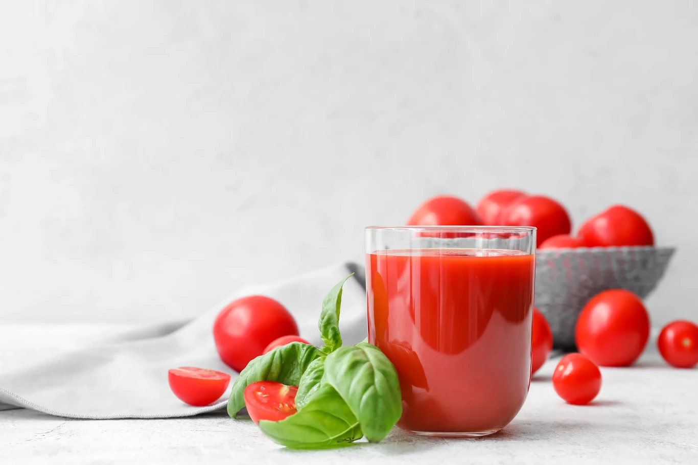 Szklanka soku pomidorowego ustawiona obok świeżych pomidorów i liści bazylii, na tle miski wypełnionej pomidorami.