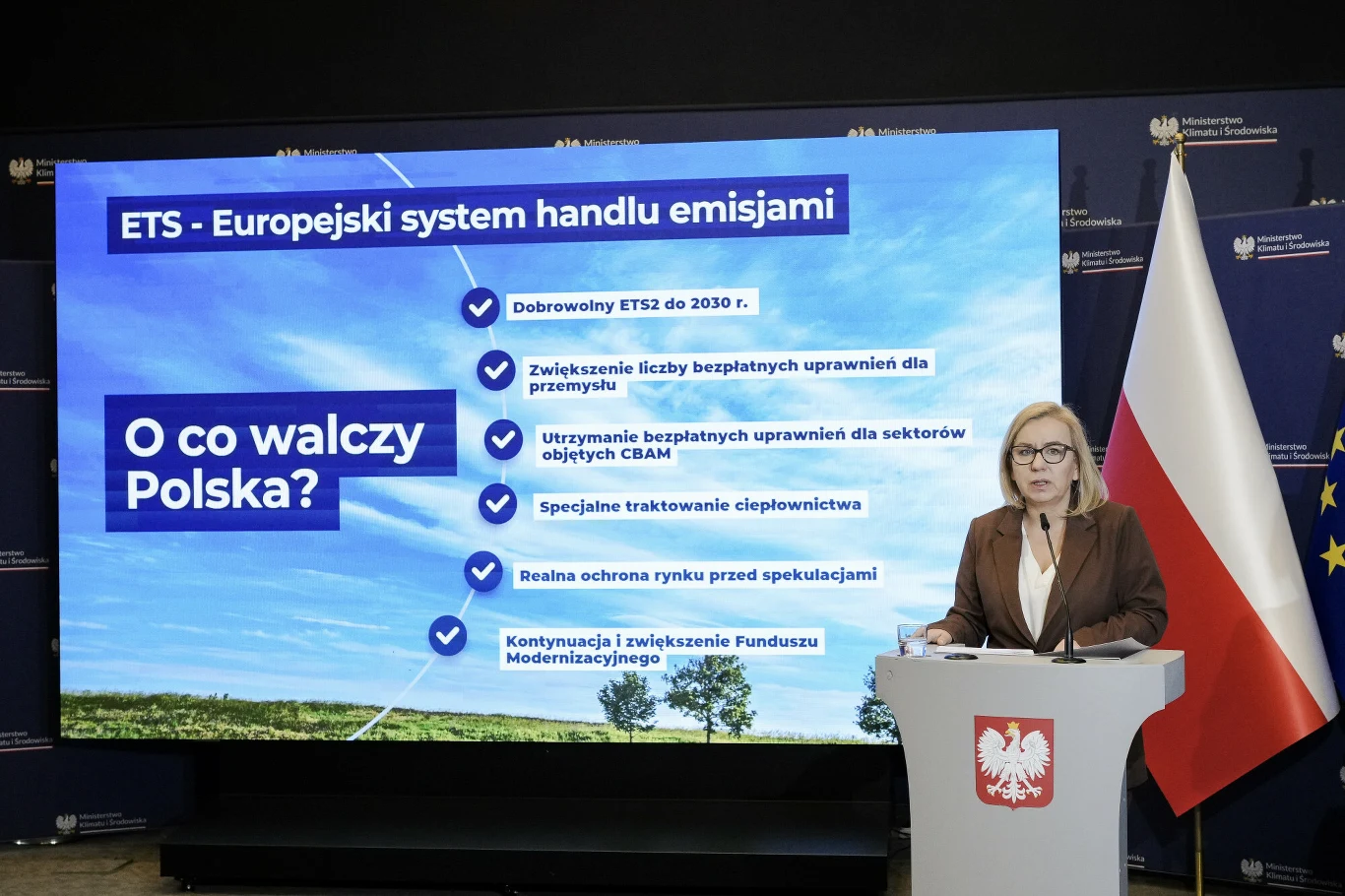 Kobieta stojąca przy mównicy z godłem Polski na tle dużego ekranu z informacjami dotyczącymi Europejskiego Systemu Handlu Emisjami, obok flaga Polski oraz Unii Europejskiej.