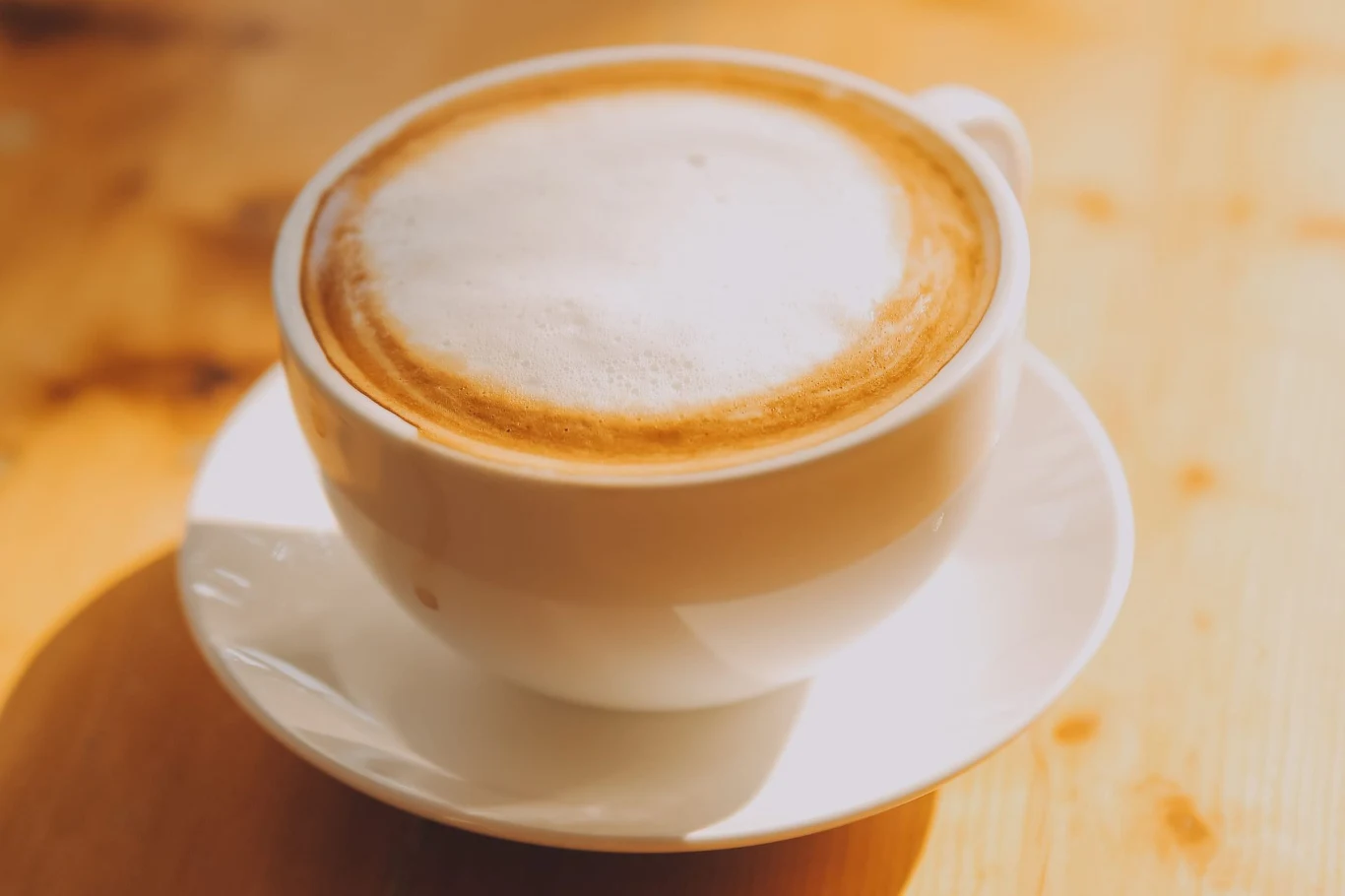 Biała filiżanka cappuccino z gęstą warstwą mlecznej pianki ustawiona na spodku na drewnianym stole w jasnym świetle dziennym.