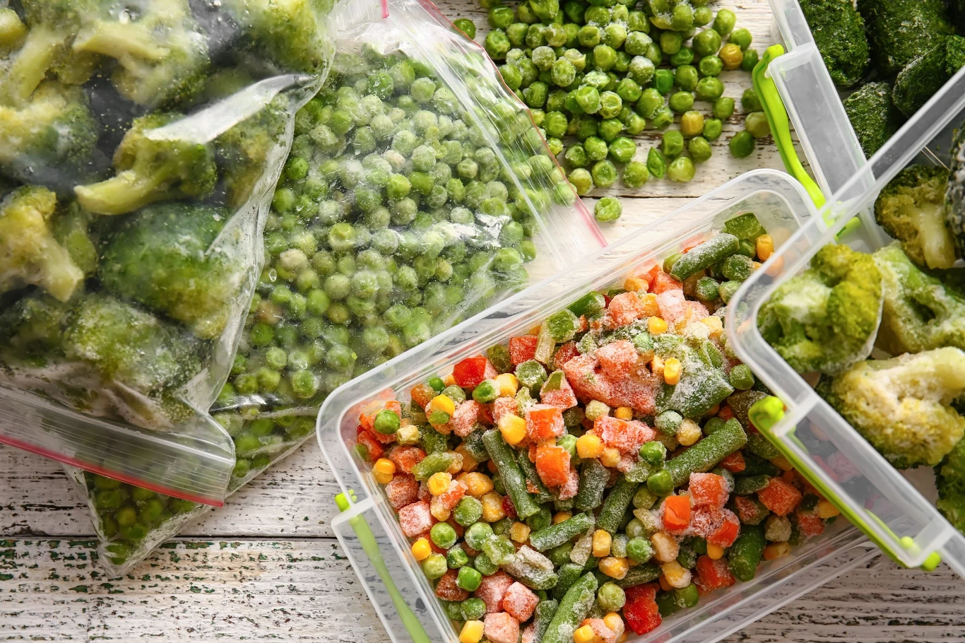 zamrożone warzywa w plastikowych pojemnikach i woreczkach, w tym brokuły, groszek oraz mieszanka warzywna z kukurydzą, marchewką i zieloną fasolką, ułożone na jasnym, drewnianym blacie