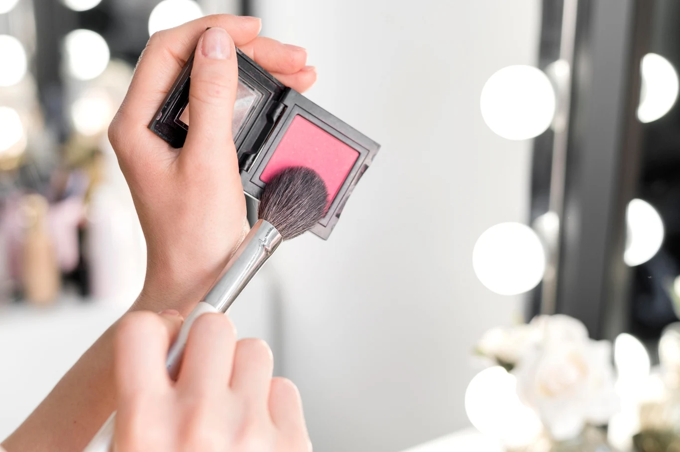 Dłoń trzymająca opakowanie różu do policzków oraz pędzel nabierający kosmetyk, z rozmytym lustrem oświetlonym żarówkami w tle.