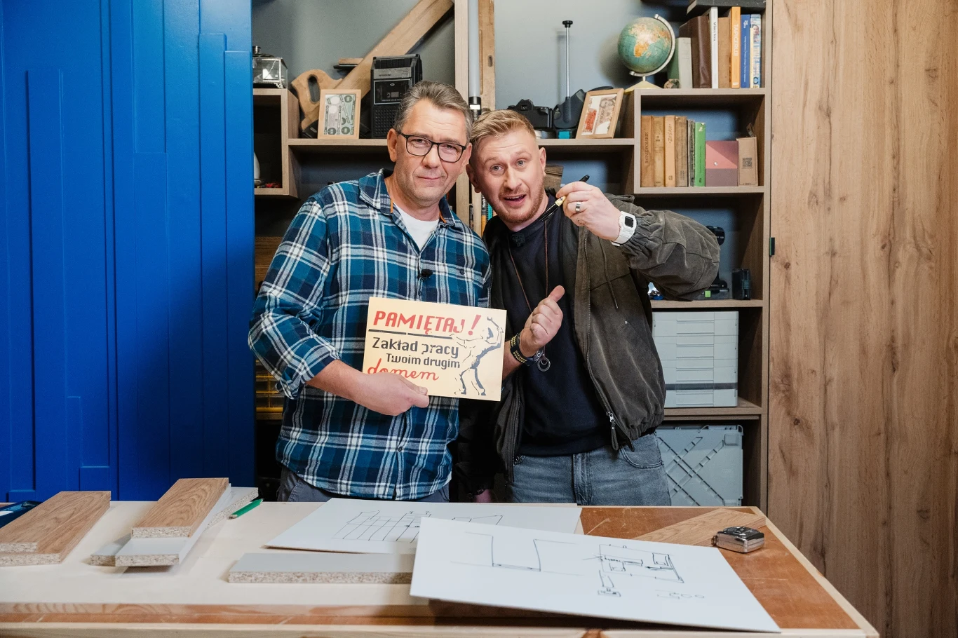 Dwóch mężczyzn stoi razem przy drewnianym stole warsztatowym, jeden z nich trzyma tabliczkę ostrzegawczą z napisem 'PAMIĘTAJ! Zakład pracy i teren drogi, zachowaj ostrożność', na stole leżą drewniane elementy oraz rysunki techniczne, w tle regał z ksią...