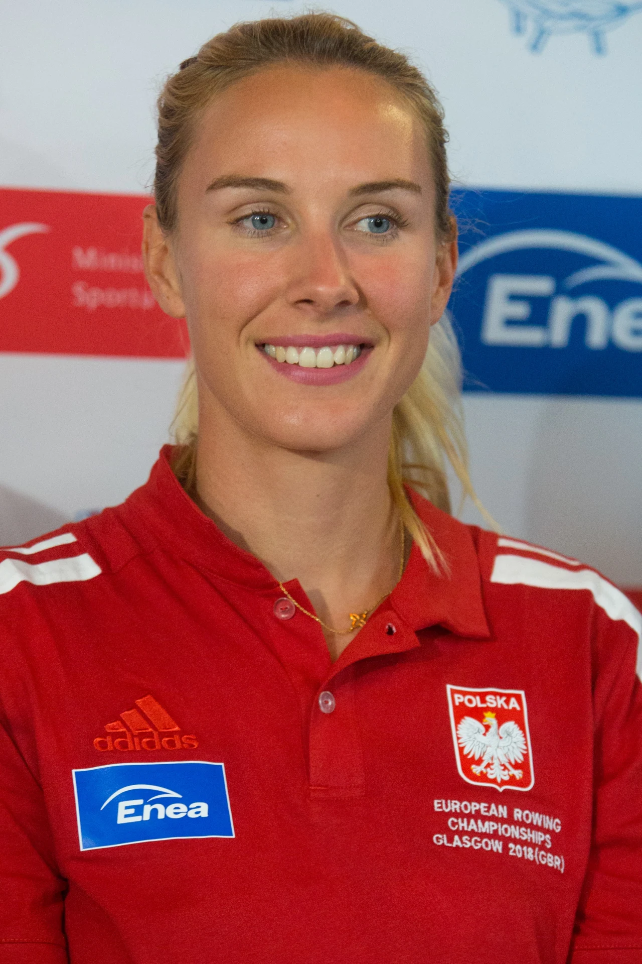 kobieta w czerwonej koszulce reprezentacji Polski z logo Adidas i Enea oraz napisem European Rowing Championships Glasgow 2018, uśmiechnięta, znajdująca się na tle z logotypami sponsorów
