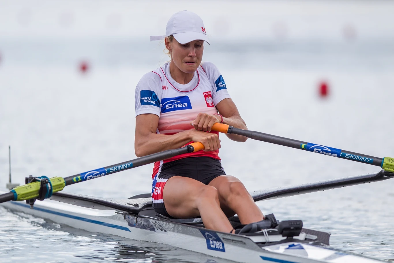 Kobieta w stroju sportowym z biało-czerwonymi barwami Polski oraz logotypami sponsorów, wiosłująca w jednoosobowej łodzi wioślarskiej na spokojnej wodzie