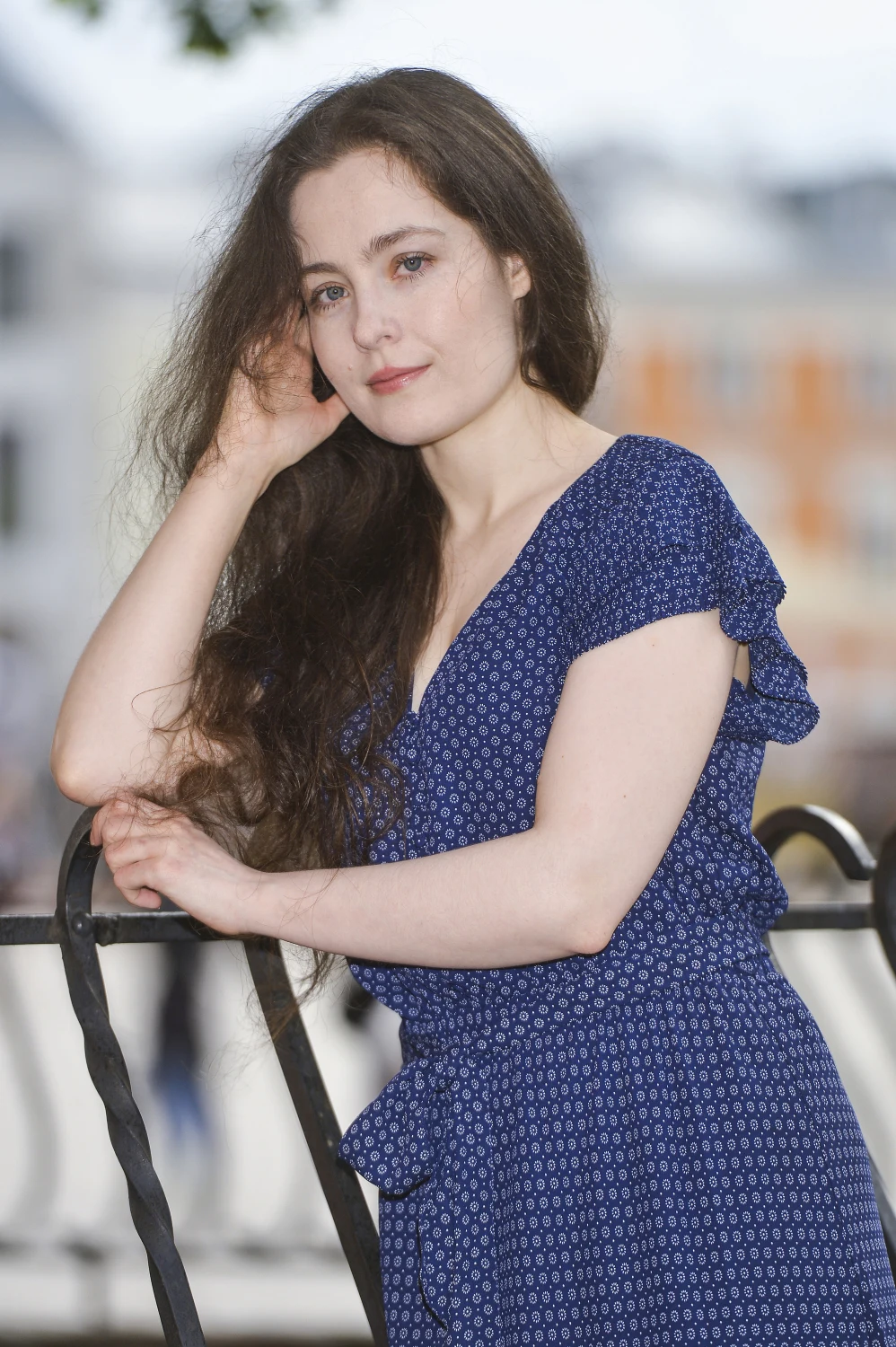 Sara Müldner w 2021 roku Młoda kobieta z długimi, ciemnymi włosami w granatowej sukience w drobny wzór opiera się o metalową balustradę, patrząc spokojnie w stronę obiektywu.