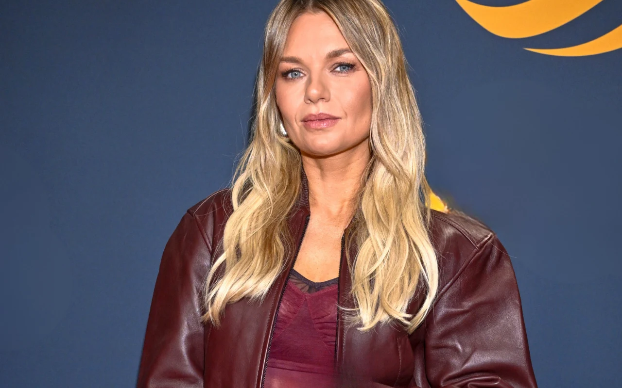 Kobieta o blond włosach ubrana w bordową kurtkę i top, stoi na tle granatowej ściany z żółtym elementem graficznym.