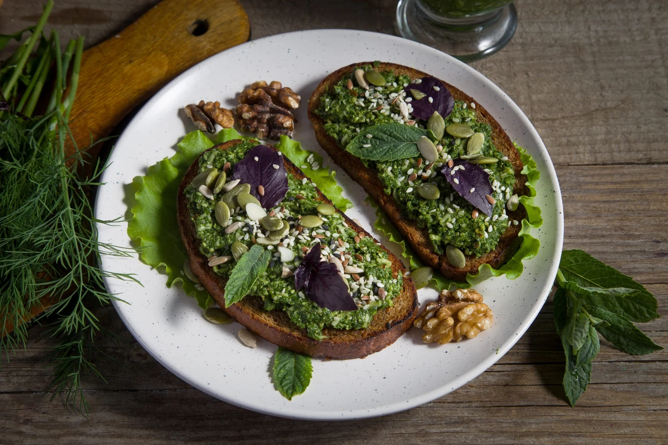 Walczysz z wiosennym przesileniem? Przygotuj tę pastę kanapkową, a odzyskasz energię. Dwie kromki chleba z pastą z zielonych warzyw, pestkami dyni i nasionami, podane na liściu sałaty obok orzechów włoskich.