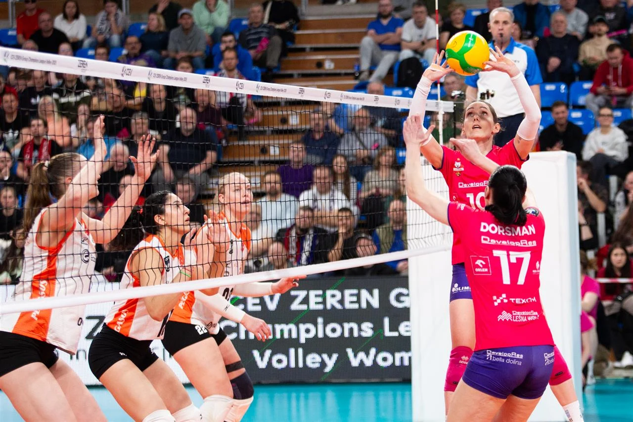 zawodniczka w różowej koszulce unosi piłkę do ataku tuż nad siatką, podczas gdy drużyna w pomarańczowych koszulkach próbuje ją zablokować, a w tle widoczna jest pełna widownia