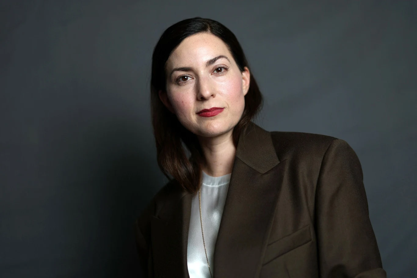 Rebecca Zlotowski Rebecca Zlotowski w brązowej marynarce i białej bluzce na szarym tle, patrzy w stronę obiektywu.