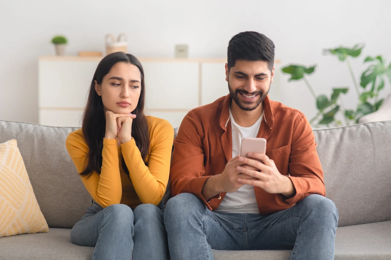 Mężczyzna siedzi na kanapie, skupiony i uśmiechnięty patrzy w telefon, podczas gdy kobieta obok wyraża niezadowolenie i zmartwienie, opierając twarz na dłoniach i spoglądając w jego stronę.