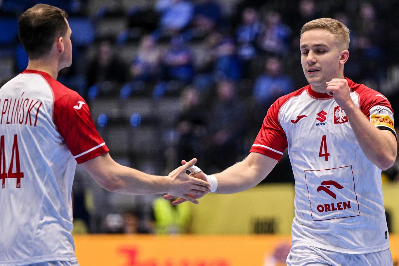 Dwóch polskich piłkarzy ręcznych w białych koszulkach z czerwonymi rękawami wykonuje gest przybicia piątki podczas meczu, na tle rozmazanej publiczności na trybunach.