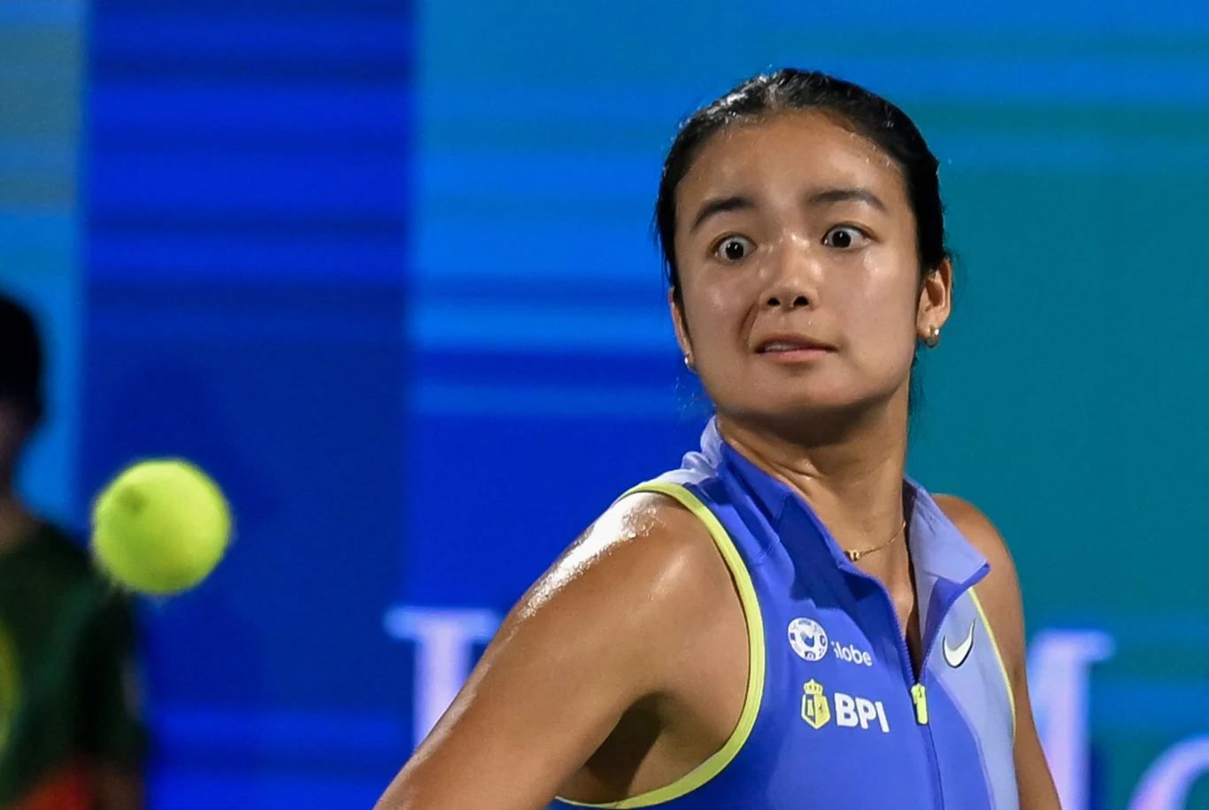 Tenisistka ubrana w niebieską koszulkę sportową z żółtymi akcentami skupia wzrok na zbliżającej się do niej piłce tenisowej, wyrażając zaskoczenie na twarzy, a w tle widać rozmyte niebieskie pasy.