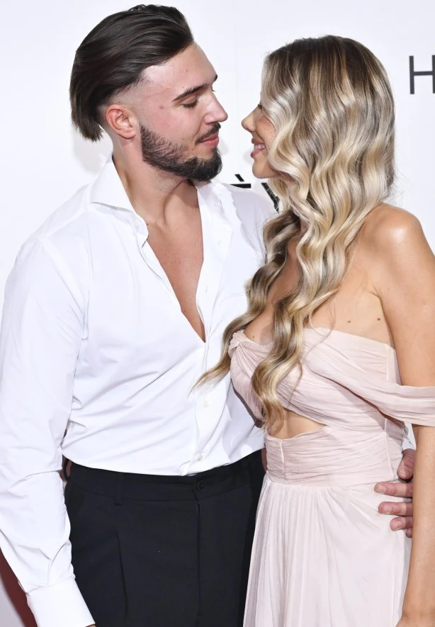 Para w eleganckich strojach patrzy sobie w oczy z uśmiechem; mężczyzna w białej, rozpiętej koszuli obejmuje kobietę w jasnoróżowej sukni z odkrytymi ramionami i długimi falowanymi włosami.