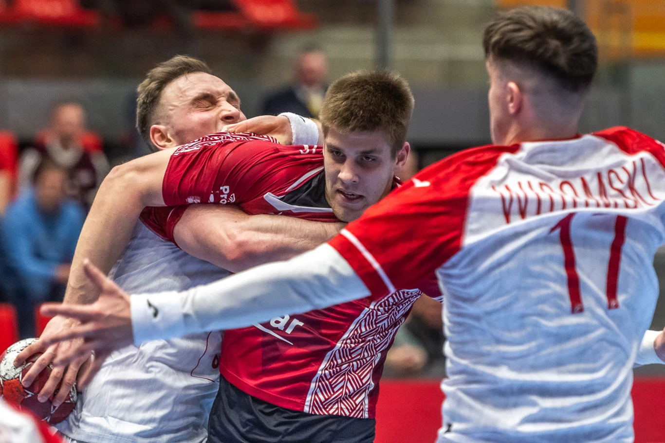 trzech zawodników walczy o piłkę podczas meczu piłki ręcznej, jeden z nich w czerwonej koszulce jest zaciśnięty w objęciach przez przeciwnika, a drugi w białej koszulce z numerem 11 próbuje zablokować ruch