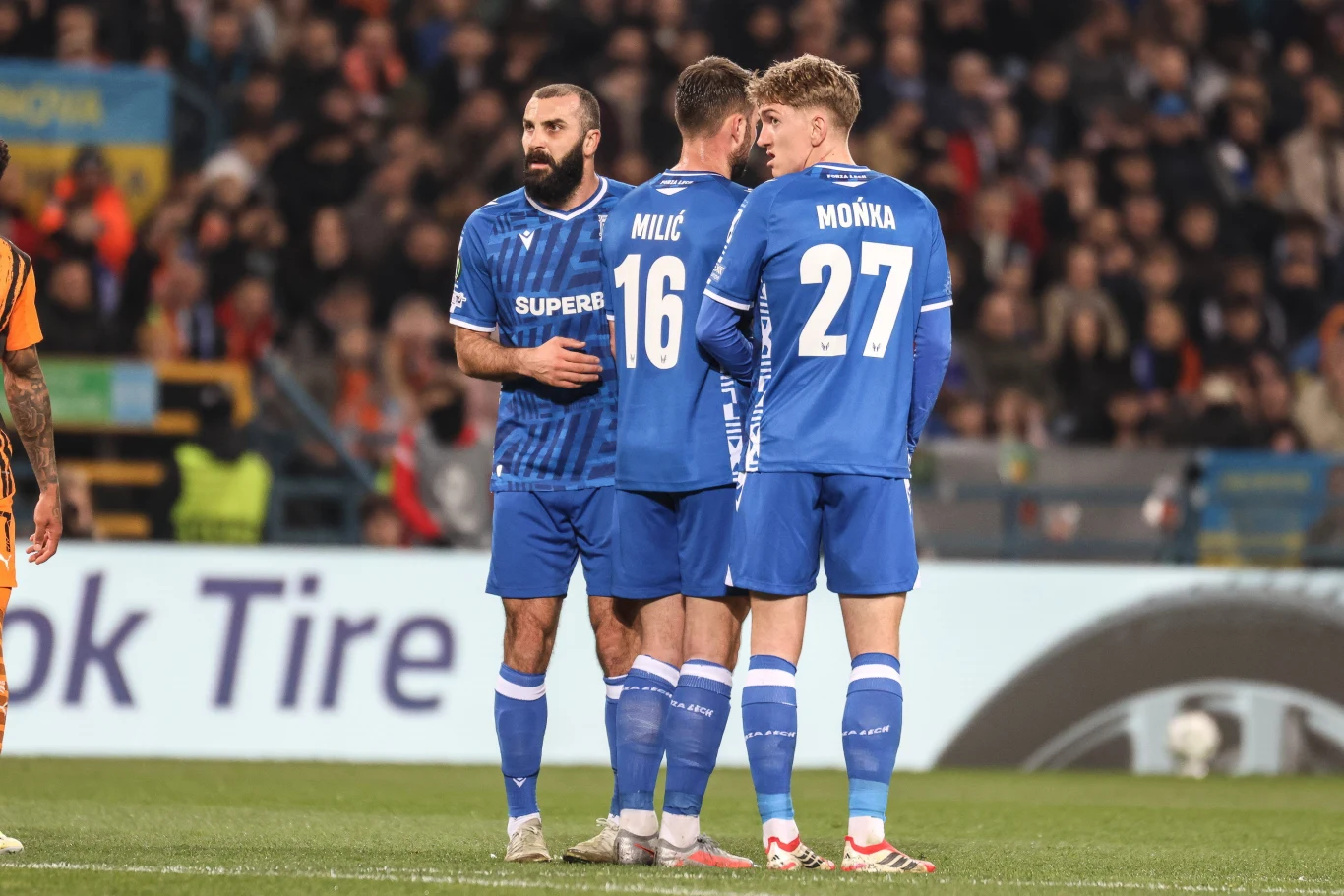 Trzech piłkarzy w niebieskich strojach drużyny ustawionych blisko siebie na murawie stadionu, jeden z nich spogląda w stronę aparatu, a w tle widoczni są kibice na trybunach.
