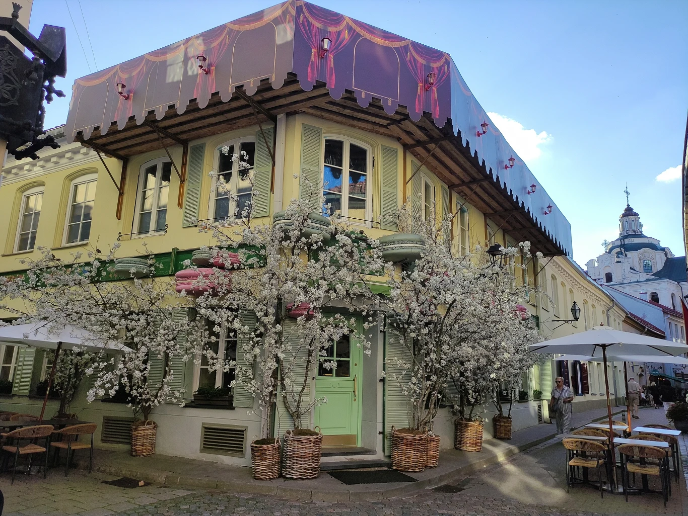 dwupiętrowa kamienica z licznymi, białymi dekoracjami kwiatowymi wokół wejścia i okien, nad wejściem kolorowy daszek imitujący zasłony, wokół ogródkowe stoliki z parasolami, w tle fragment zabytkowego kościoła