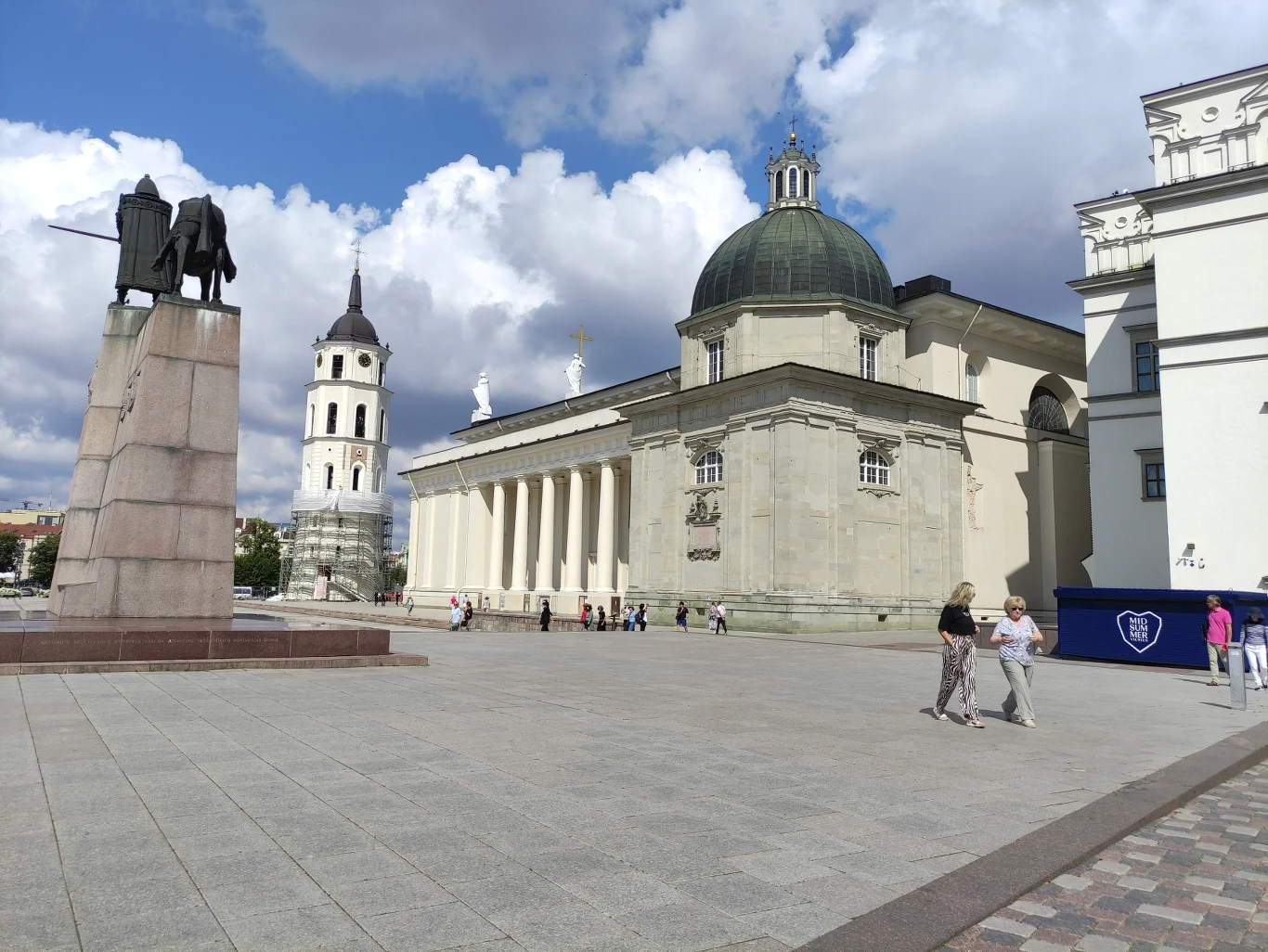 Plac z widoczną katedrą o jasnej fasadzie i zielonej kopule, obok wysoka biała dzwonnica oraz pomnik z postaciami na podwyższeniu, wokół przechadzają się ludzie przy pięknej pogodzie z niebem pełnym chmur.