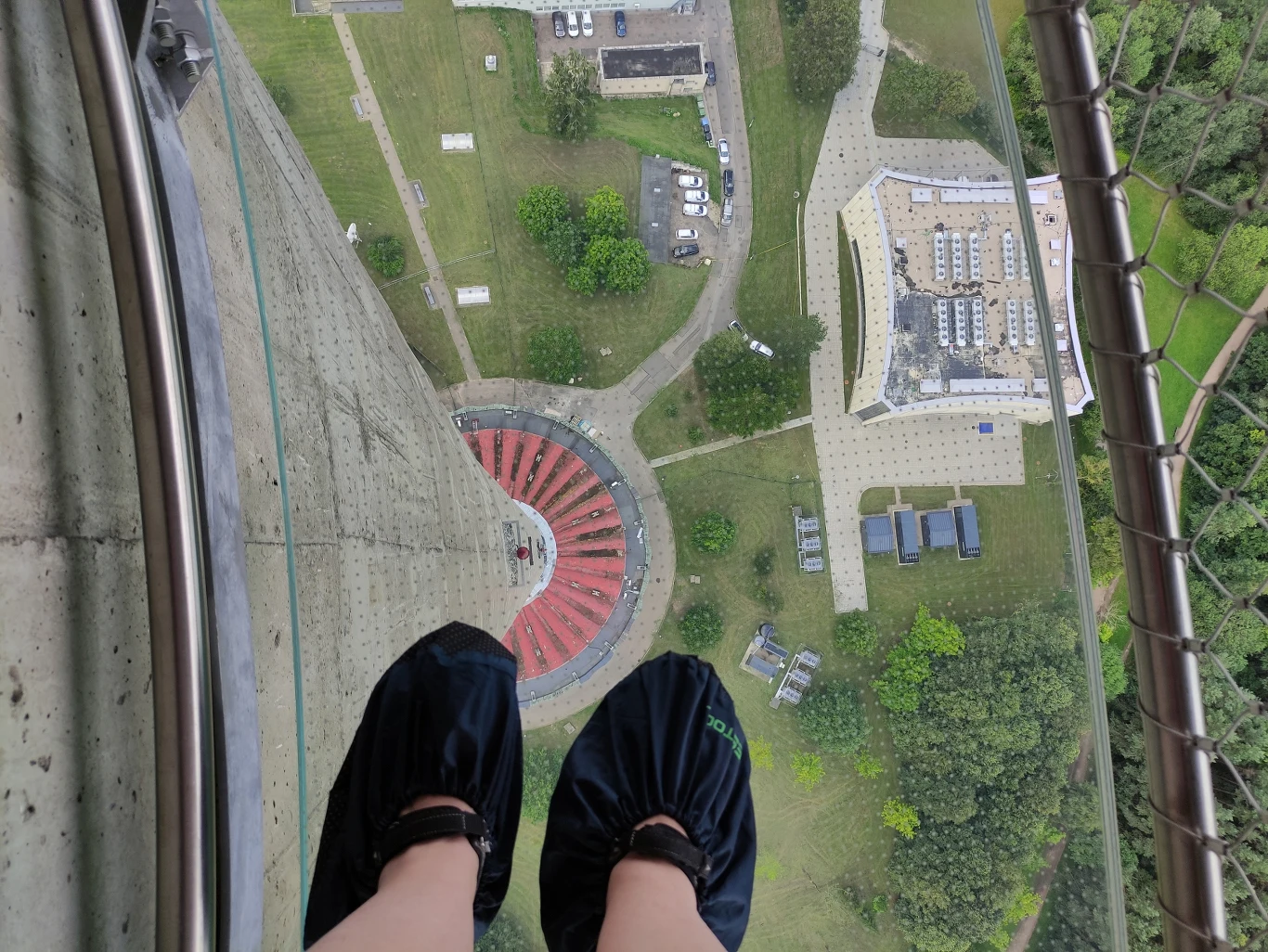 Stopy w ochraniaczach stojące na przezroczystej podłodze wieży widokowej wysoko nad ziemią, z widokiem na plac, zielone trawniki, drzewa oraz otaczające budynki.