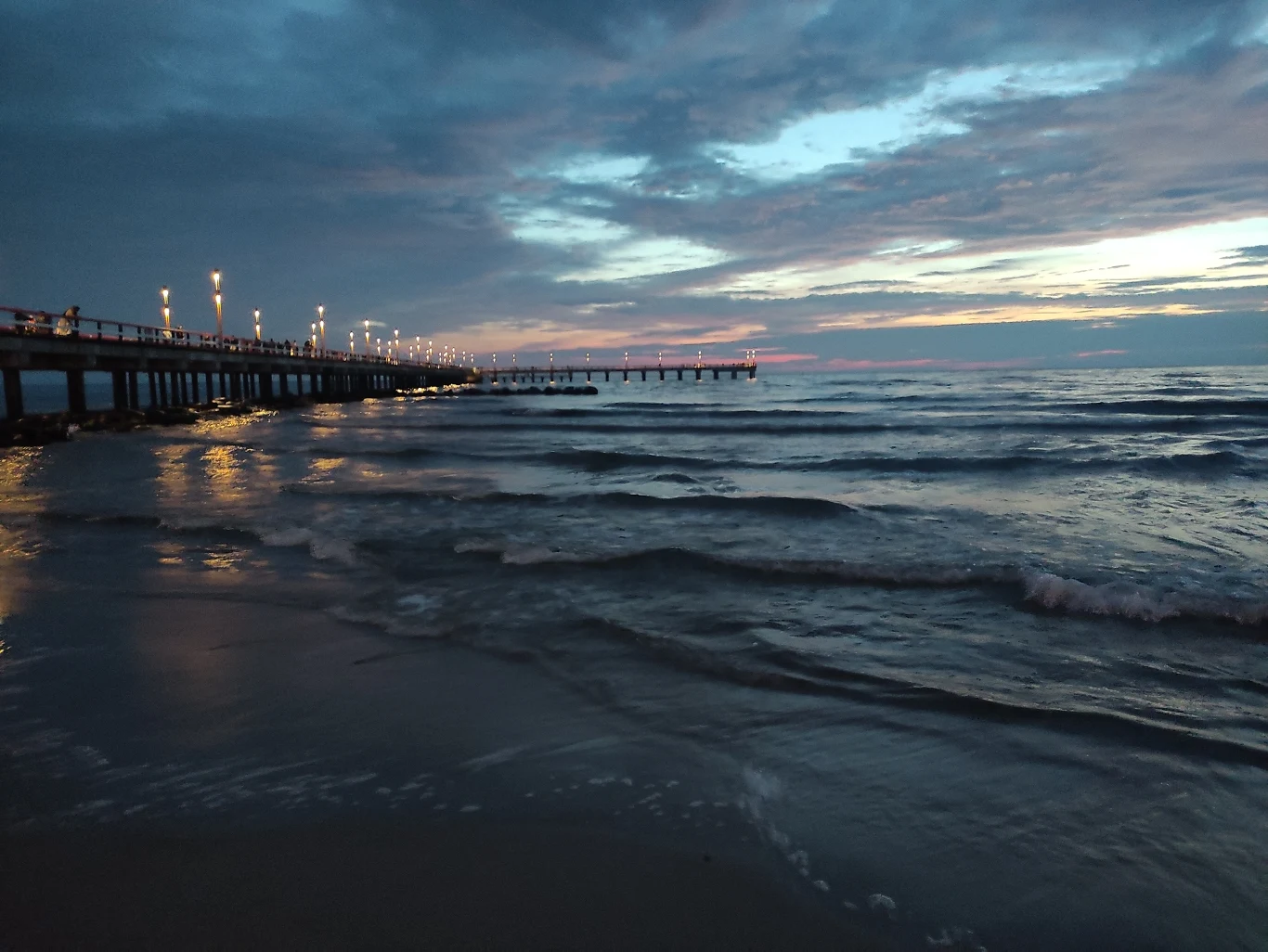 molo oświetlone latarniami ciągnące się nad wzburzonym morzem podczas zmierzchu, niebo z chmurami i subtelnymi barwami zachodu słońca odbijającymi się na powierzchni wody