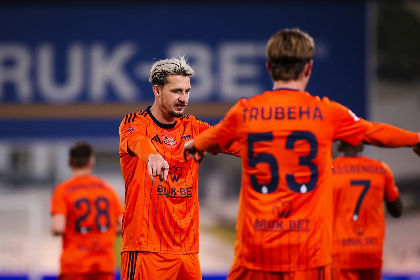 piłkarze drużyny w pomarańczowych strojach podczas meczu, zawodnik z przodu wskazuje kierunek lub wydaje polecenia koledze z numerem 53 na plecach