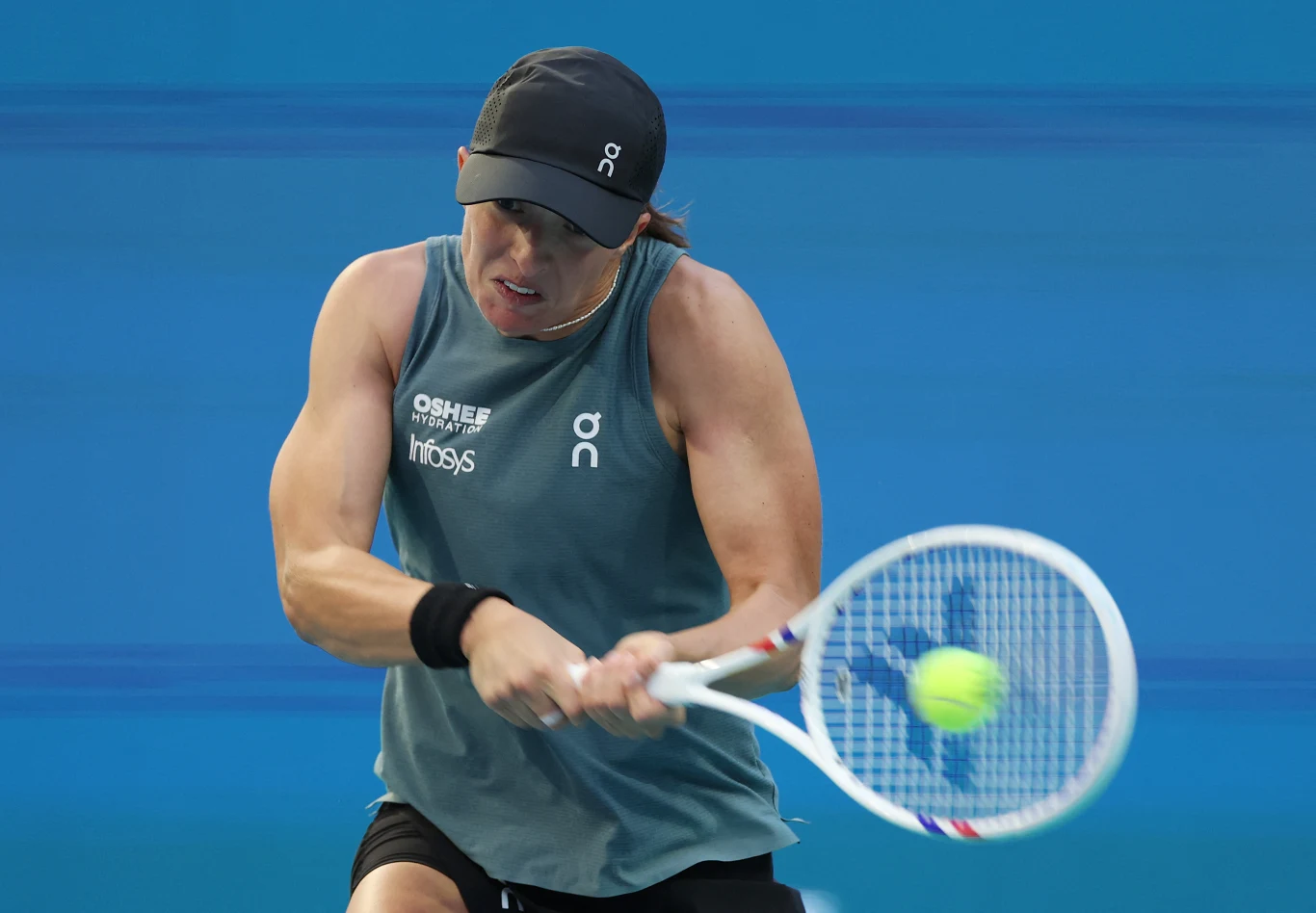 tenisistka w czapce i sportowej koszulce wykonuje mocny backhand dwuręczny, skupiona na odbiciu piłki na tle niebieskiego kortu