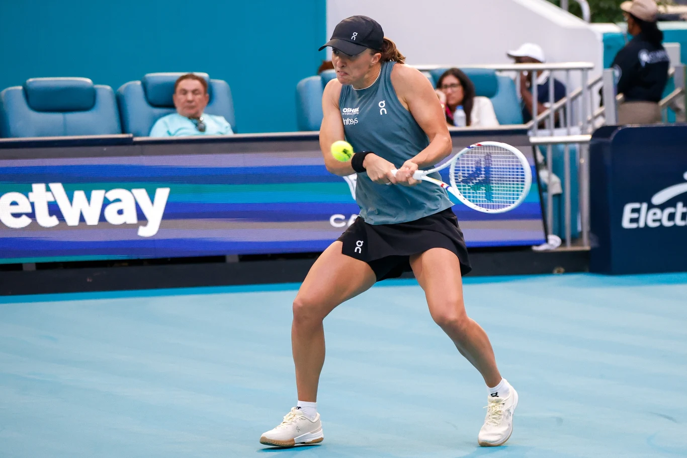 tenisistka w niebieskim stroju wykonuje uderzenie forhendowe na profesjonalnym korcie, skupiona na piłce, wokół widownia i reklamy sponsorów