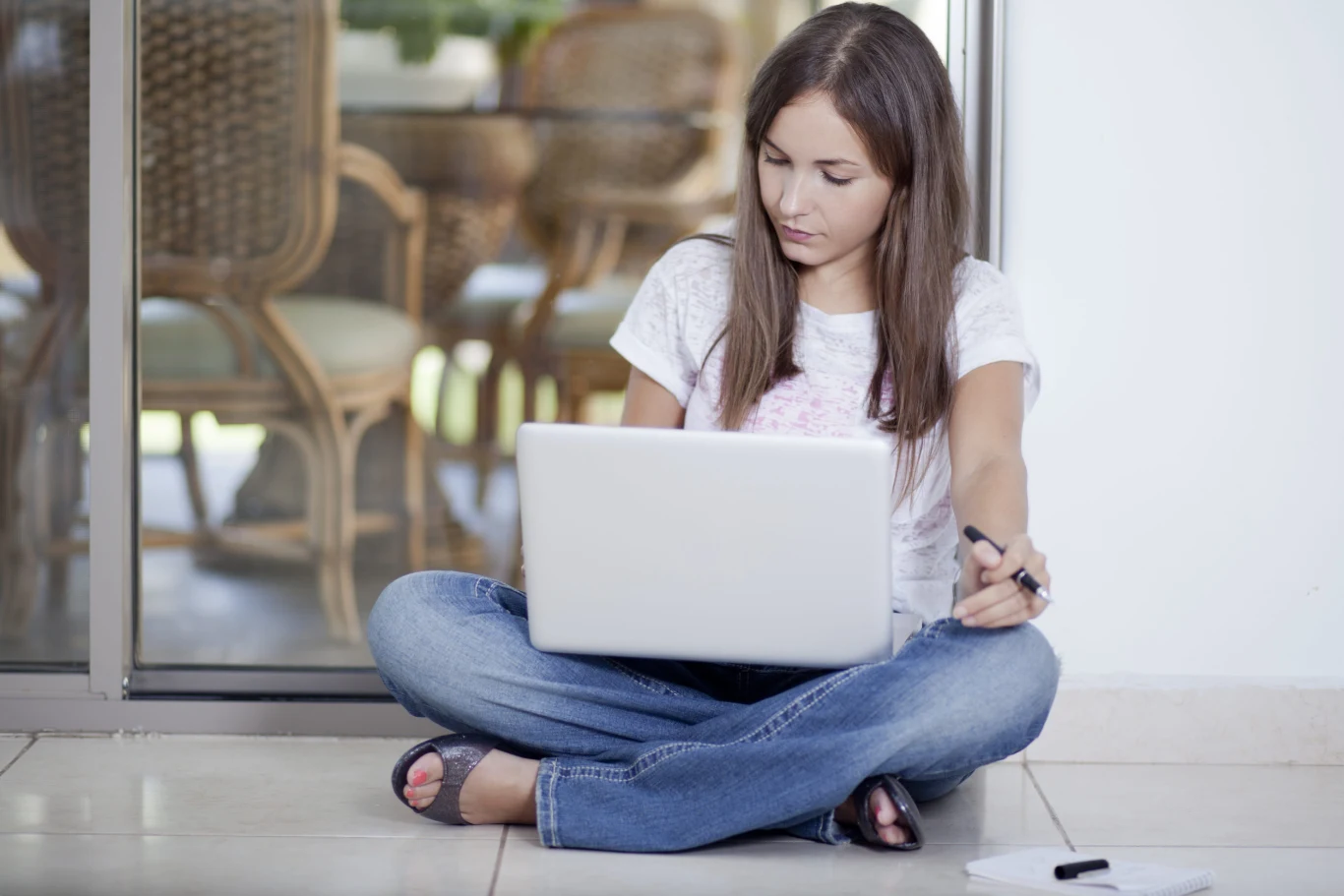 Młoda kobieta w dżinsach i białej koszulce siedzi po turecku na podłodze, pracując przy laptopie; w jednej ręce trzyma długopis, a obok leży notatnik.