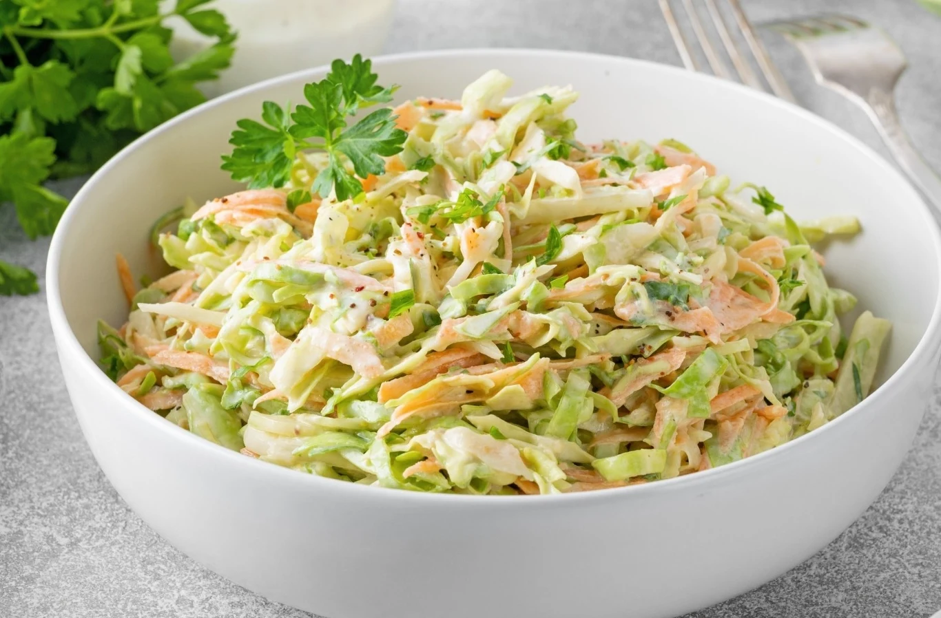 Jak zrobić pyszną i zdrową surówkę? Poznaj prosty przepis Miska surówki z kapusty, marchewki i pietruszki w kremowym sosie, udekorowana pietruszką.