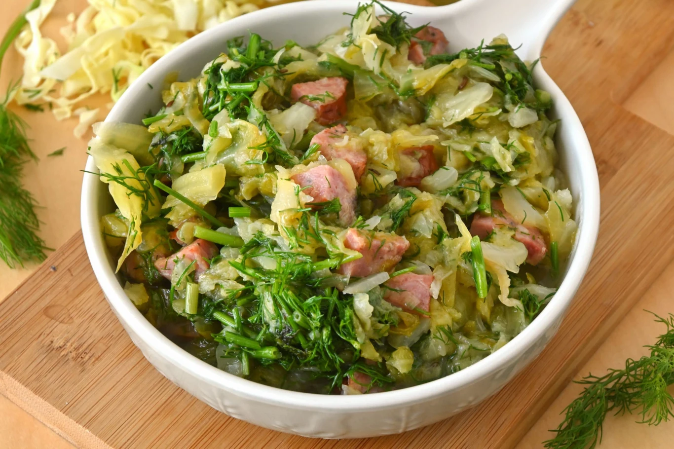 Kapusta pekińska na ciepło to pyszny dodatek do obiadu Misa z duszoną kapustą pekińską, kawałkami kiełbasy i koperkiem, podana na drewnianej desce.