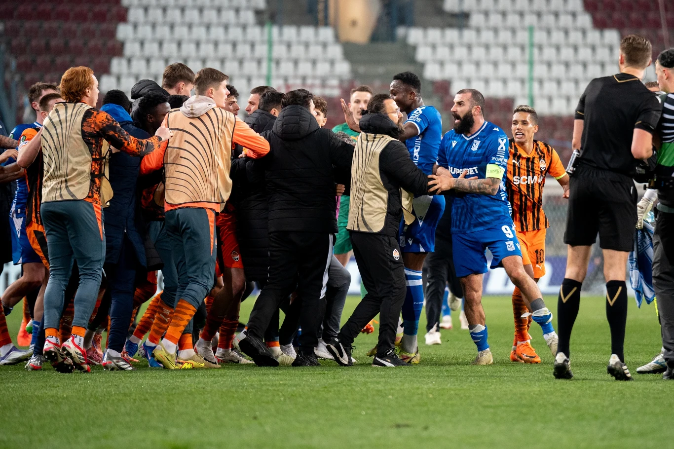 grupa piłkarzy w mieszanych strojach sportowych w trakcie dynamicznej przepychanki na murawie stadionu, otoczeni przez kilku arbitrów próbujących opanować sytuację
