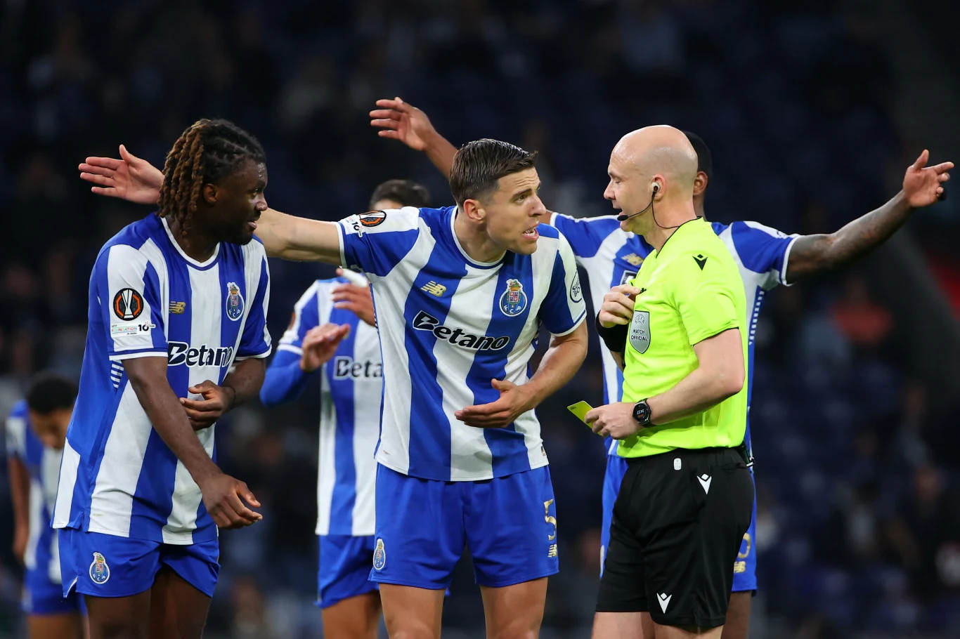 Zawodnicy drużyny w biało-niebieskich strojach energicznie dyskutują z arbitrem ubranym na czarno, wyrażając niezadowolenie z decyzji podczas meczu piłkarskiego.