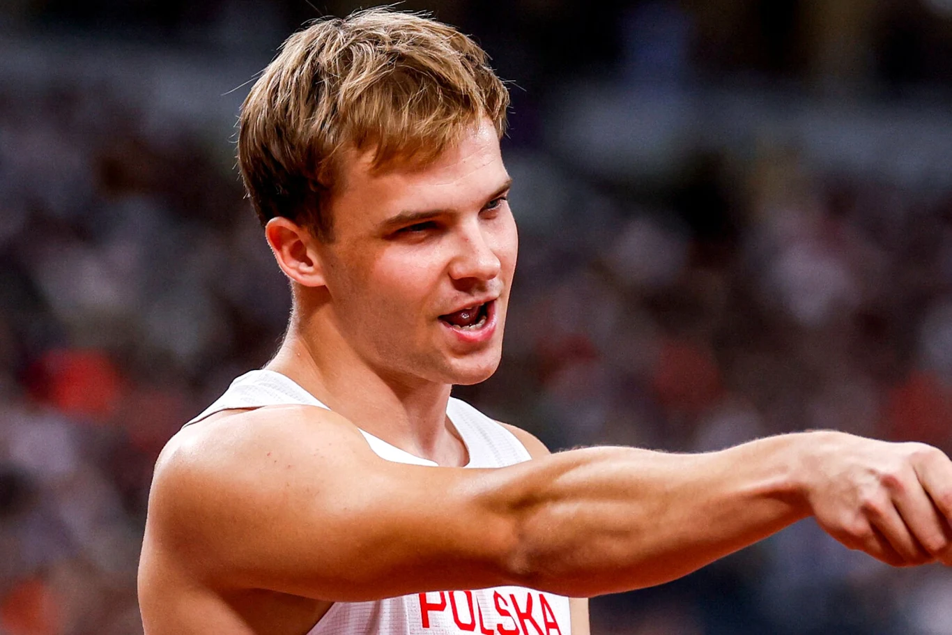 Młody sportowiec w białej koszulce z napisem 'Polska' energicznie gestykuluje w trakcie zawodów lekkoatletycznych, w tle niewyraźna publiczność.