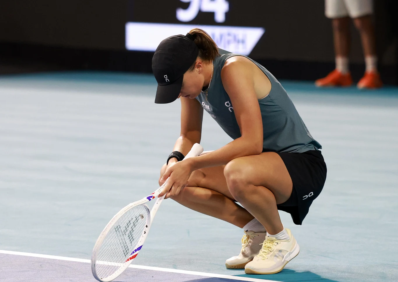 Iga Świątek kuca na korcie tenisowym w sportowym stroju i czapce, trzyma rakietę opartą o ziemię