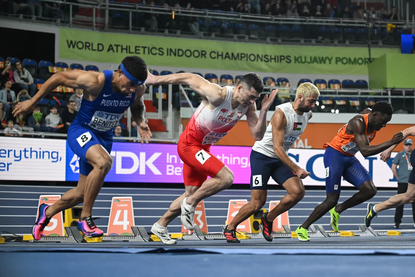 Grupa sprinterów w różnych kolorach strojów startuje równocześnie z bloków na bieżni podczas halowych mistrzostw świata w lekkiej atletyce.