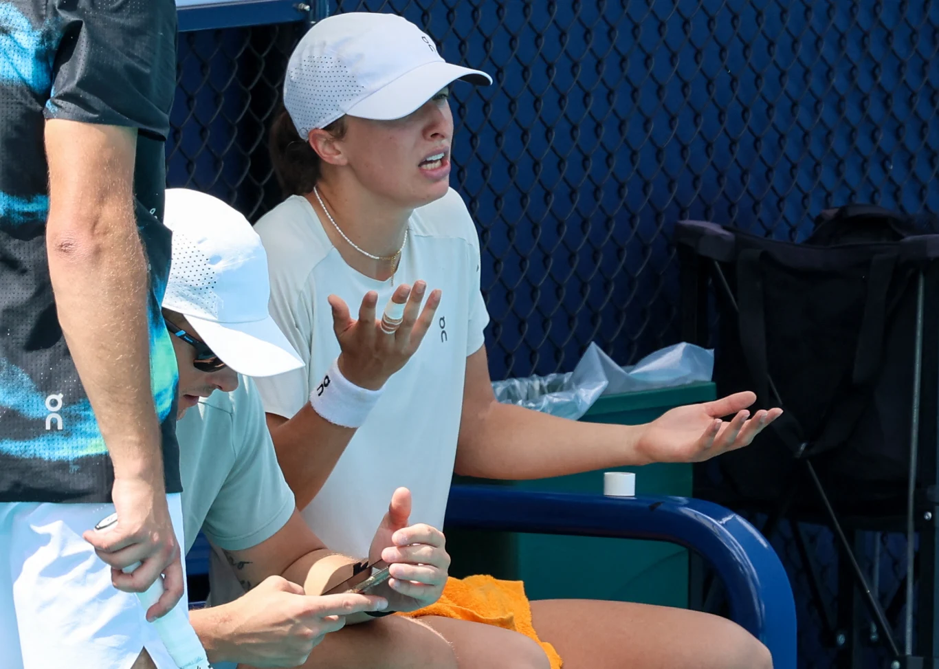 Tenisistka w białej czapce i koszulce siedzi na ławce, trzymając dłonie w geście zdziwienia lub frustracji, obok niej siedzi osoba w podobnym stroju pochylona nad smartfonem lub tabletem; w tle siatka kortu tenisowego.