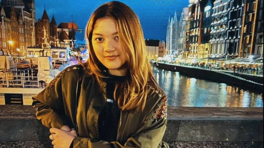 Młoda kobieta z długimi, jasnobrązowymi włosami stoi przy nabrzeżu; za nią rozświetlone nocą kamienice oraz odbijająca światła woda kanału, tworzące malowniczy, miejski krajobraz.