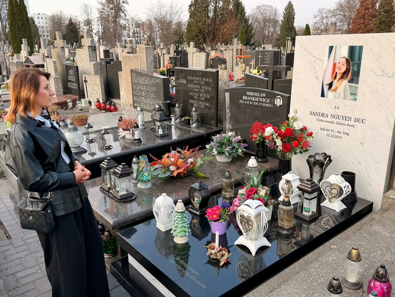 kobieta stojąca przy nowoczesnym nagrobku ozdobionym licznymi kwiatami, zniczami oraz zdjęciem młodej kobiety na płycie nagrobnej, w tle rzędy innych grobów cmentarnych i drzewa bez liści