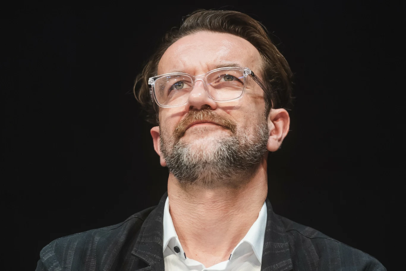 Tomasz Kot Tomasz Kot z zarostem i w okularach, w białej koszuli i ciemnej marynarce na czarnym tle.