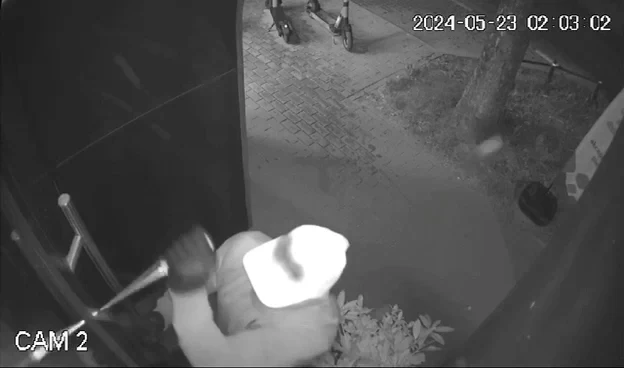 Osoba w jasnej czapce i rękawiczkach próbuje siłą otworzyć drzwi przy użyciu narzędzia, na zewnątrz widoczny chodnik, drzewo oraz dwa rowery dziecięce przywiązane do ogrodzenia.