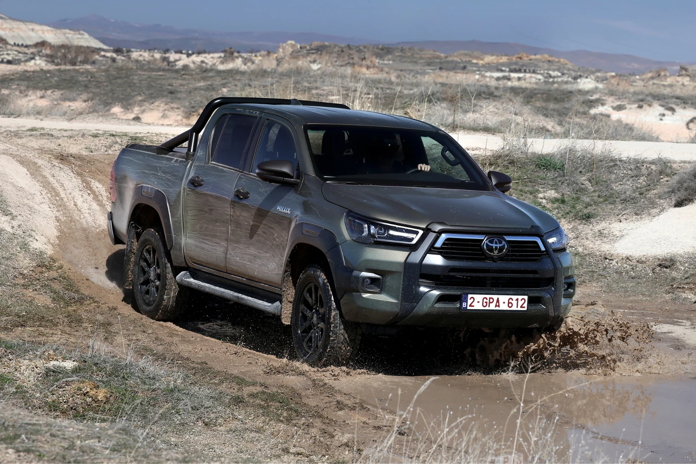 szary pick-up Toyota Hilux pokonuje błotnistą drogę terenową, unosząc wokół siebie wodę i błoto, otoczony suchym, kamienistym krajobrazem