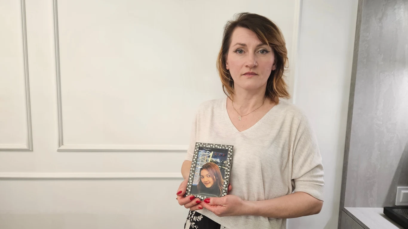 Kobieta o jasnej cerze i brązowych włosach stoi przodem do obiektywu, w dłoniach trzyma oprawione zdjęcie młodej kobiety z długimi włosami i szerokim uśmiechem.