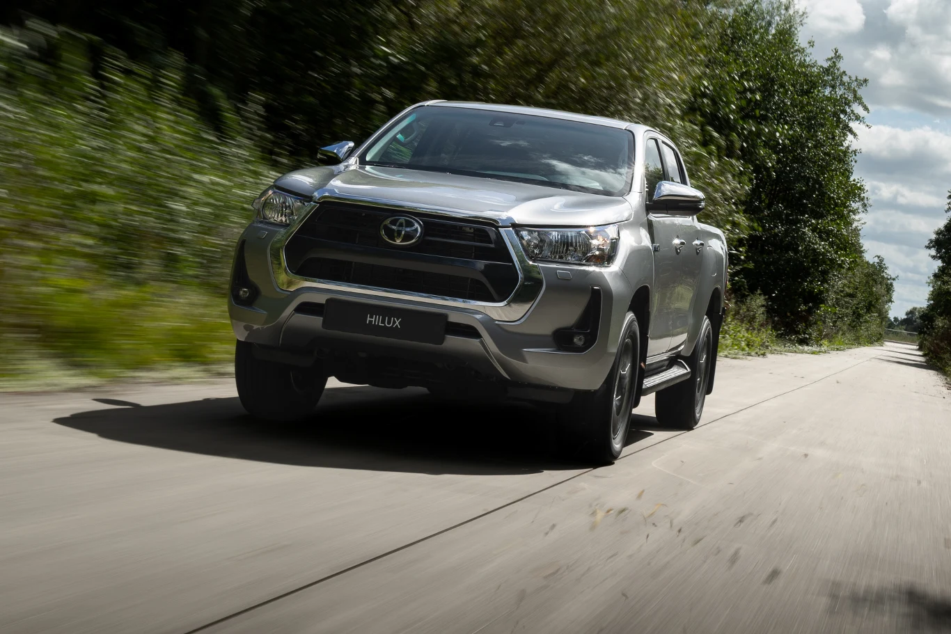 srebrny pick-up Toyota Hilux jedzie po asfaltowej drodze otoczonej zielenią, promienie słoneczne odbijają się od karoserii pojazdu, tło jest lekko rozmyte w ruchu