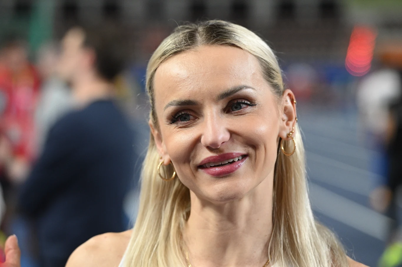 Kobieta o długich blond włosach z uśmiechem na twarzy, ubrana w sportowy strój i złote kolczyki, stoi na stadionie lekkoatletycznym, w tle rozmazane sylwetki osób i bieżnia.