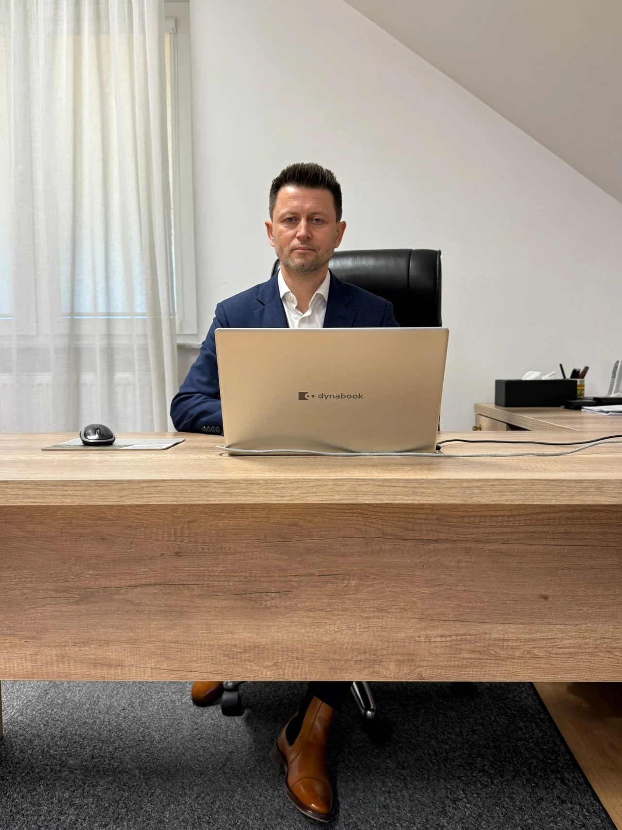 Mężczyzna w garniturze siedzi za dużym drewnianym biurkiem, pracując przy otwartym laptopie, z profesjonalnym wyrazem twarzy, w nowoczesnym, jasnym biurze.