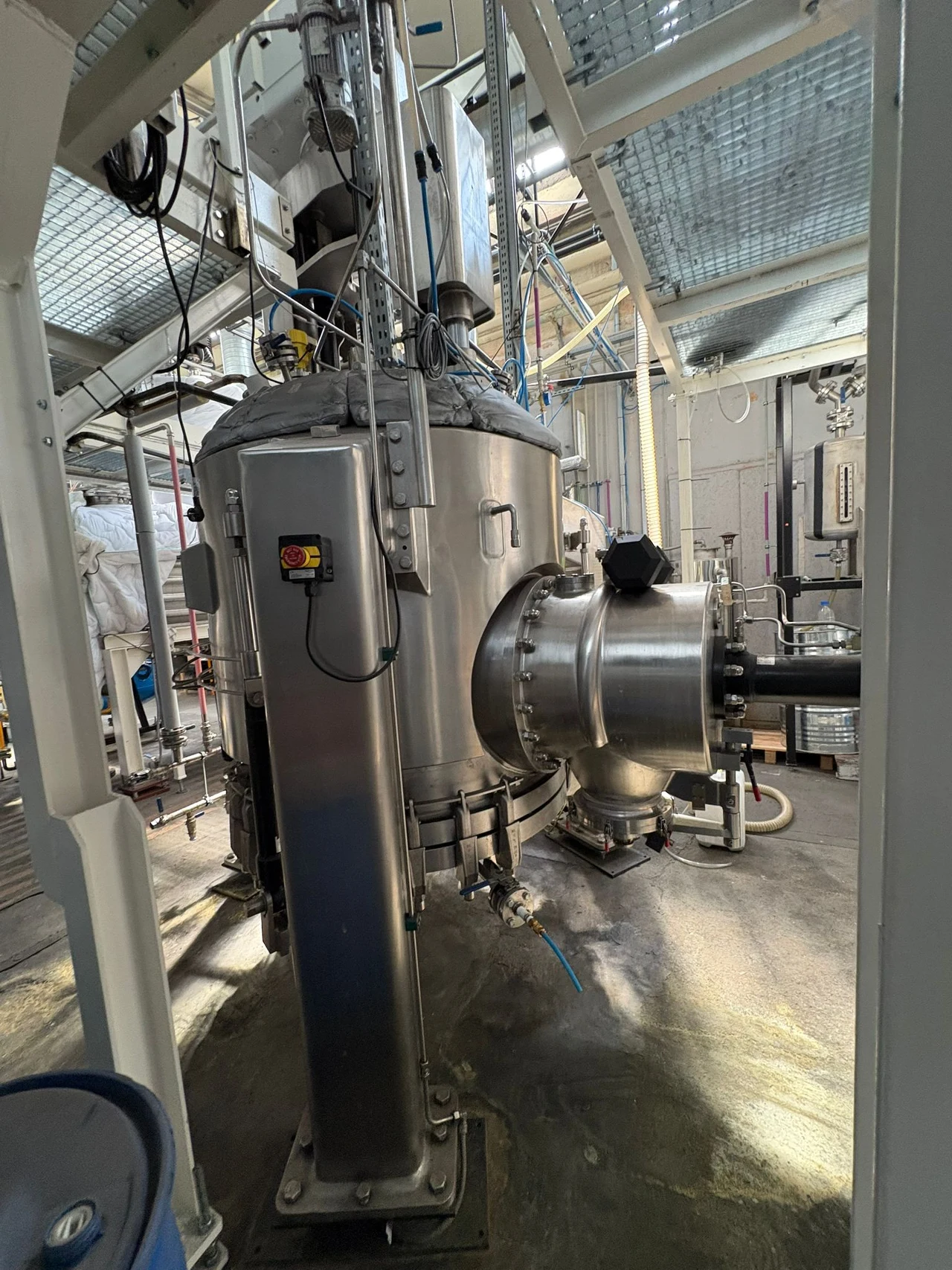 Duży przemysłowy reaktor ze stali nierdzewnej z wieloma przyłączami, zaworami oraz przewodami, osadzony na metalowej konstrukcji w nowoczesnym zakładzie produkcyjnym, otoczony instalacją technologiczną.