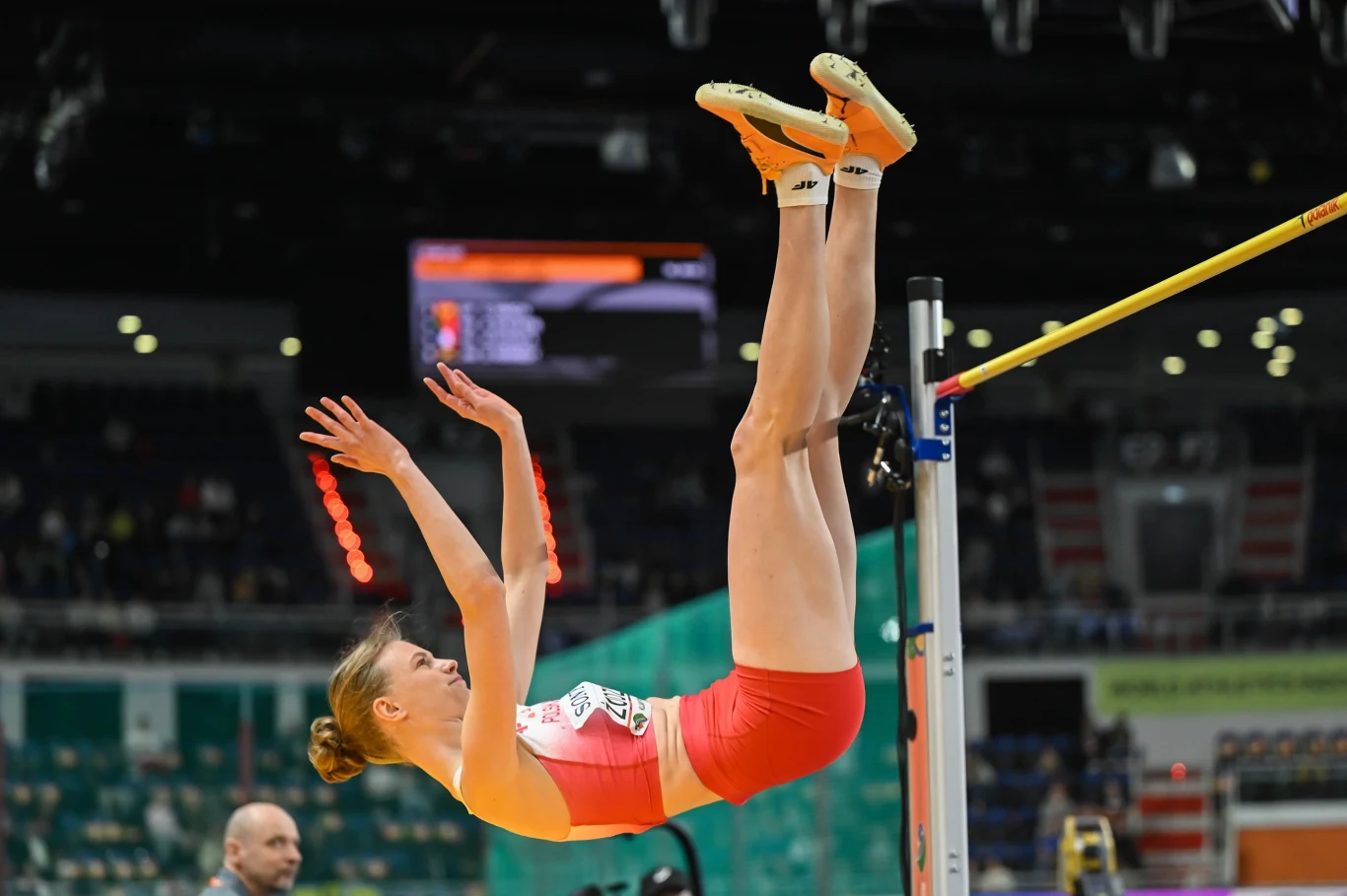 lekkoatletka w czerwonych spodenkach i białym topie wykonuje skok wzwyż, unosząc ciało nad poprzeczką podczas zawodów na stadionie lekkoatletycznym
