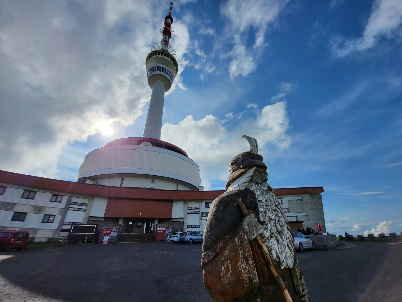 nowoczesna wieża telewizyjna górująca nad budynkiem z okrągłą podstawą, na pierwszym planie drewniana rzeźba ptaka z wysuniętym dziobem, a po bokach zaparkowane samochody i pochmurne niebo w tle
